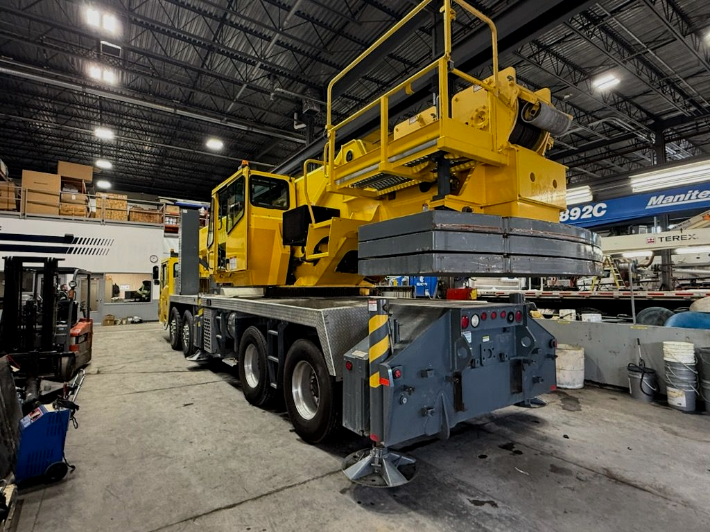Une grue mobile jaune est stationnée à l'intérieur d'un entrepôt, ses stabilisateurs déployés et des contrepoids empilés sur sa plateforme arrière.