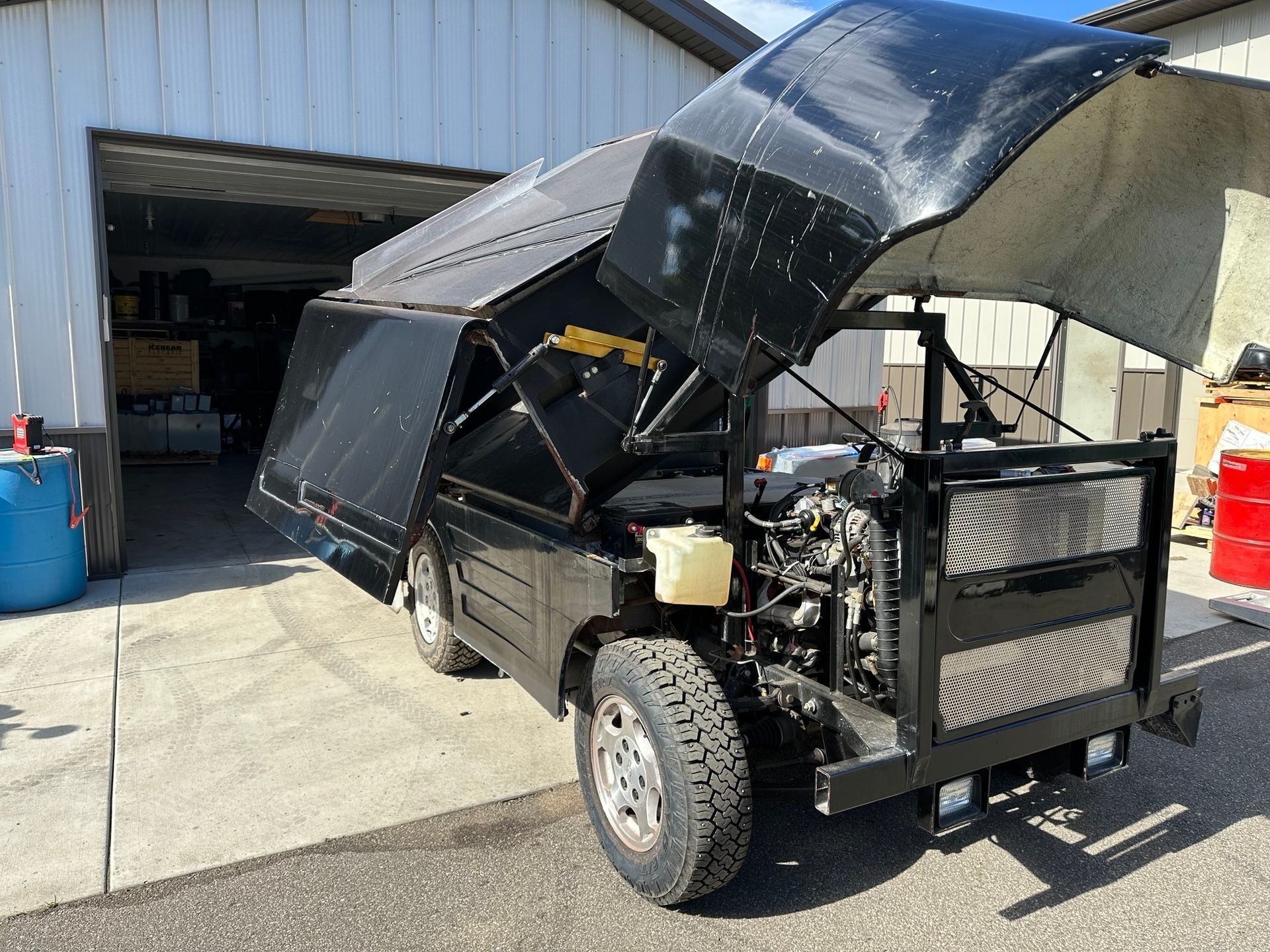 A black vehicle with the hood up is parked in front of a garage.