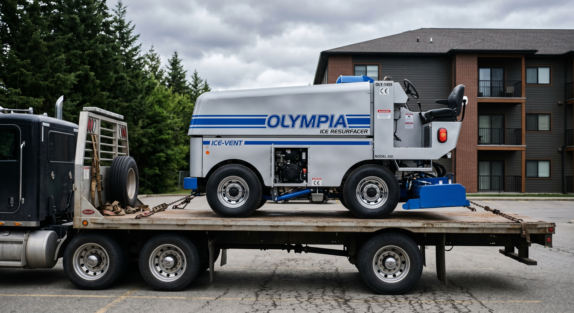 White Olympia machine on a flatbed trailer in a parking lot, with an apartment building in the background