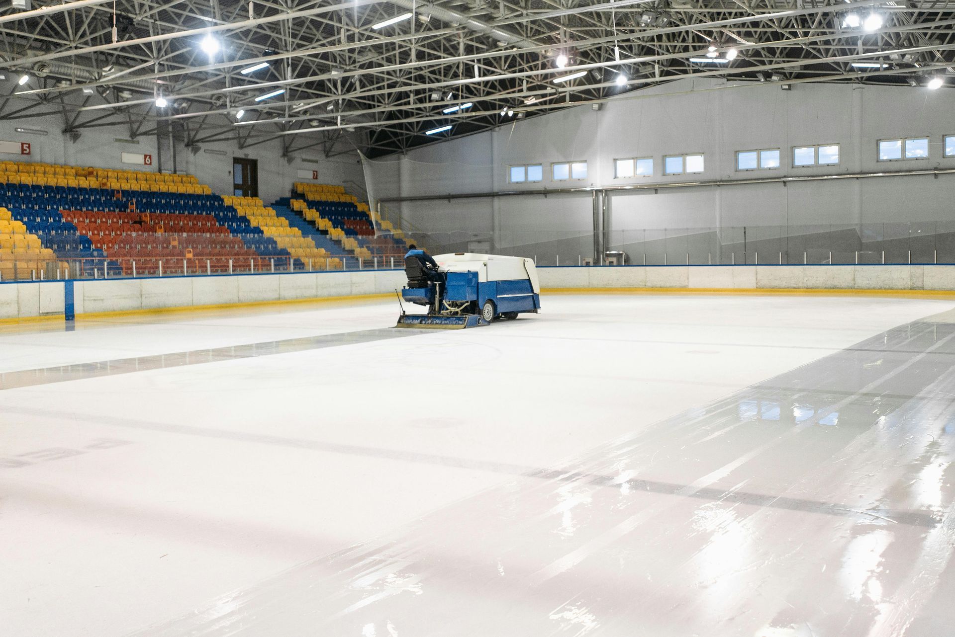 An ice resurfacing machine cleaning a large indoor ice rink with rows of colorful bleachers in the background.