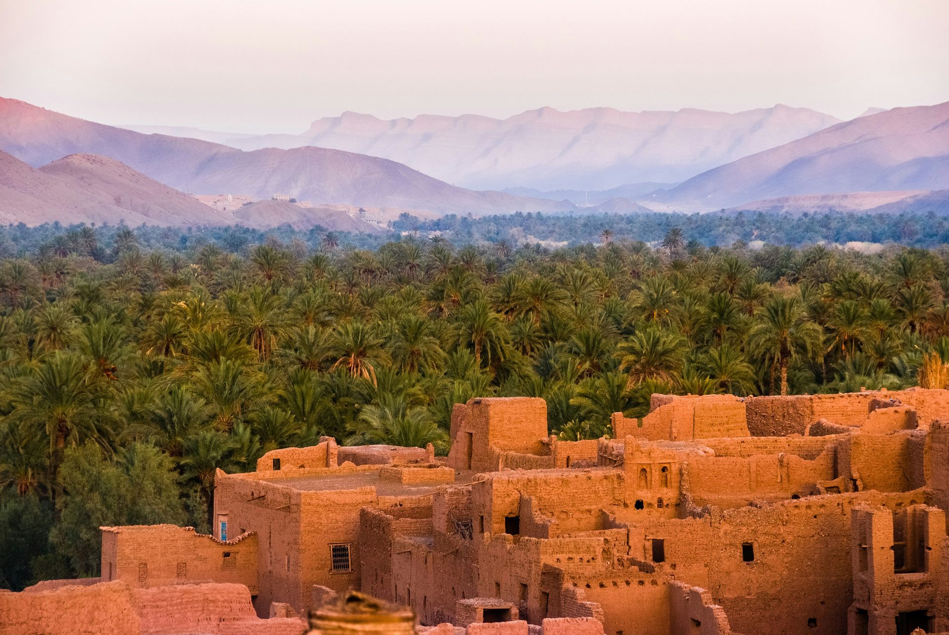 An aerial view of a desert village with palm trees and mountains in the background.