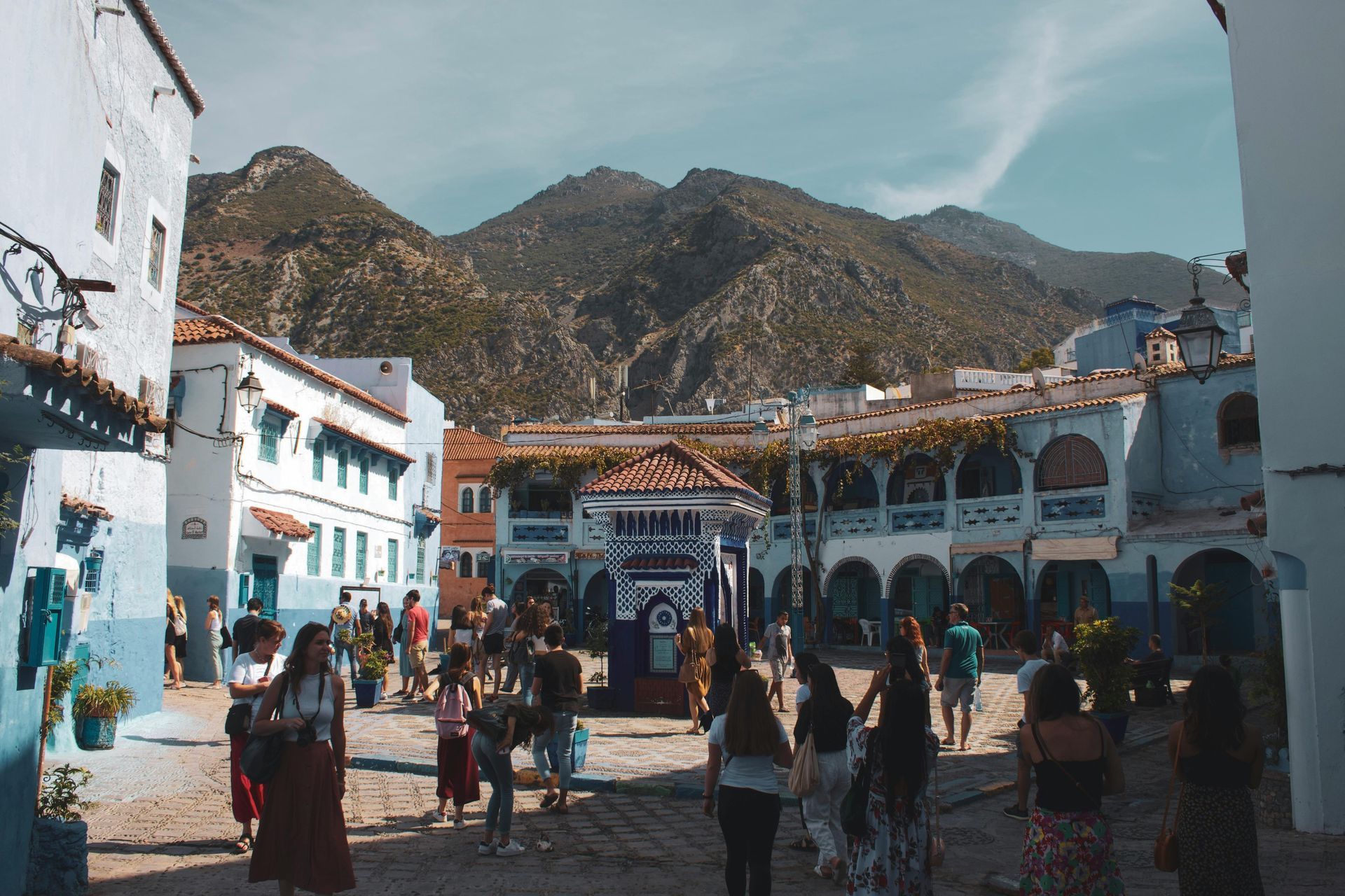 A group of people are walking down a street in a city with mountains in the background.