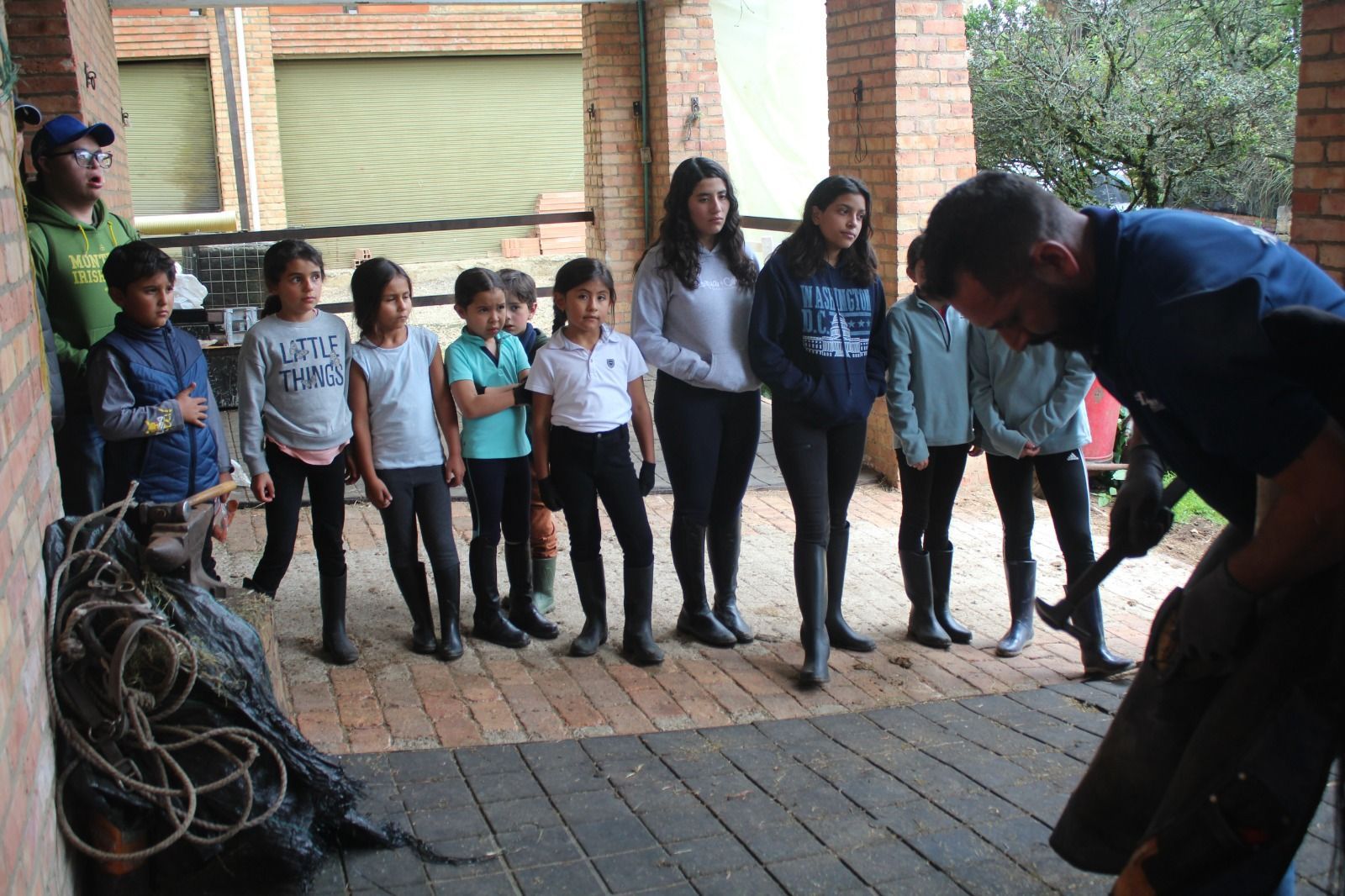 A group of children are standing in a circle watching a man shoe a horse.
