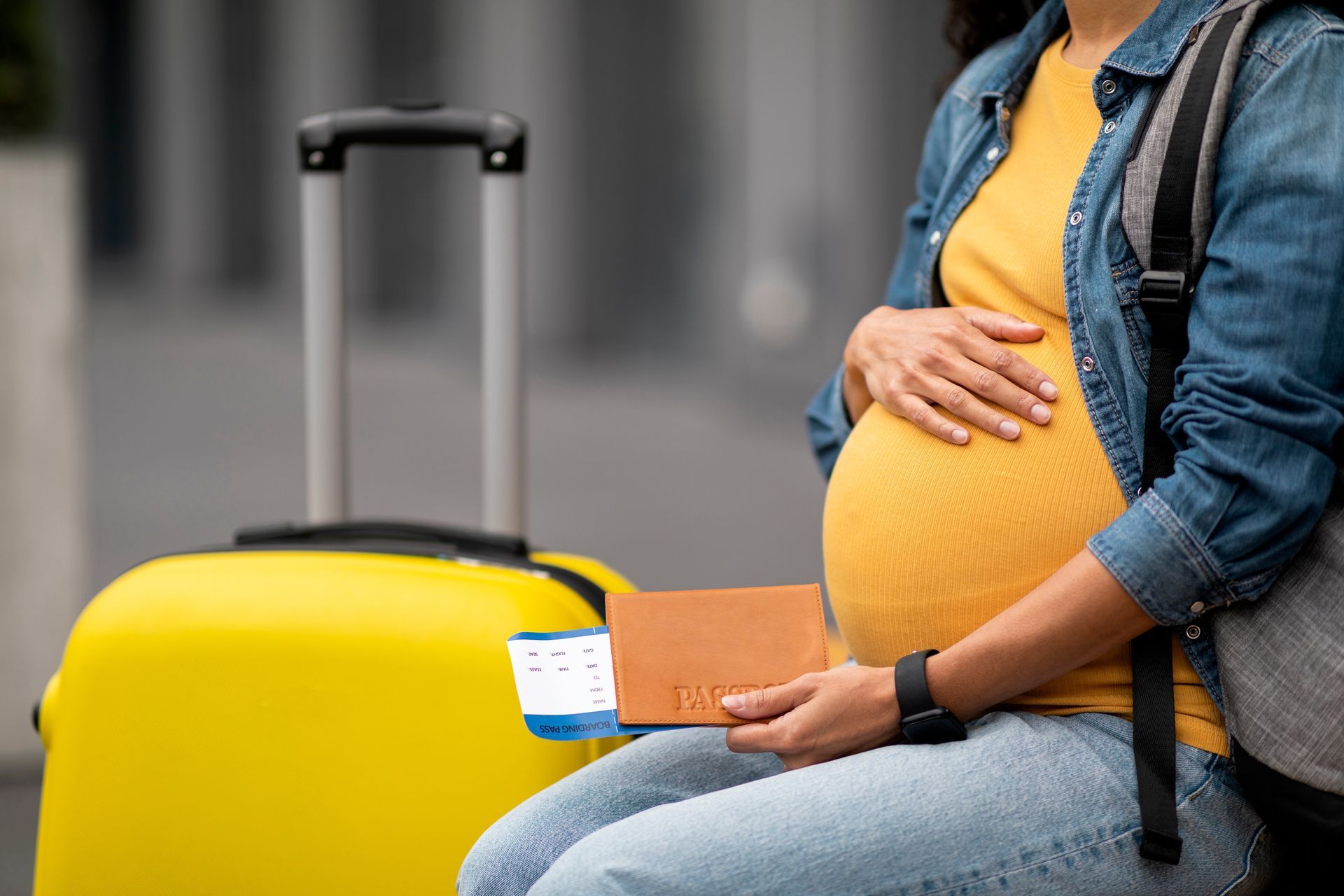 A pregnant woman is sitting next to a yellow suitcase and holding a passport.