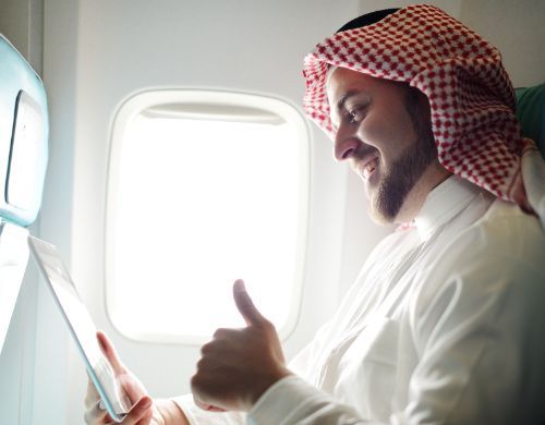 A man is sitting on an airplane looking at a tablet and giving a thumbs up.