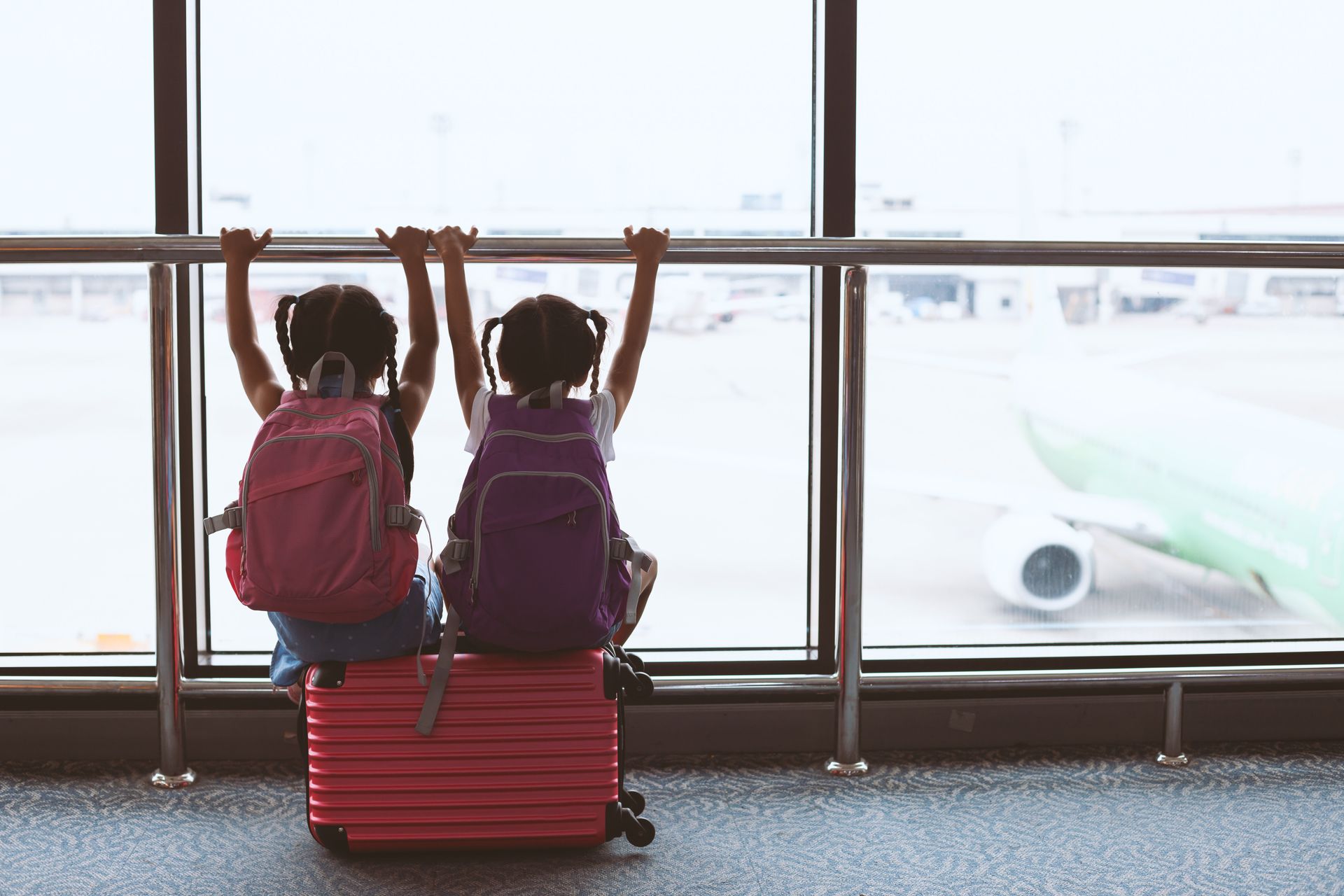 Two little girls are sitting on a suitcase looking out a window at an airport.