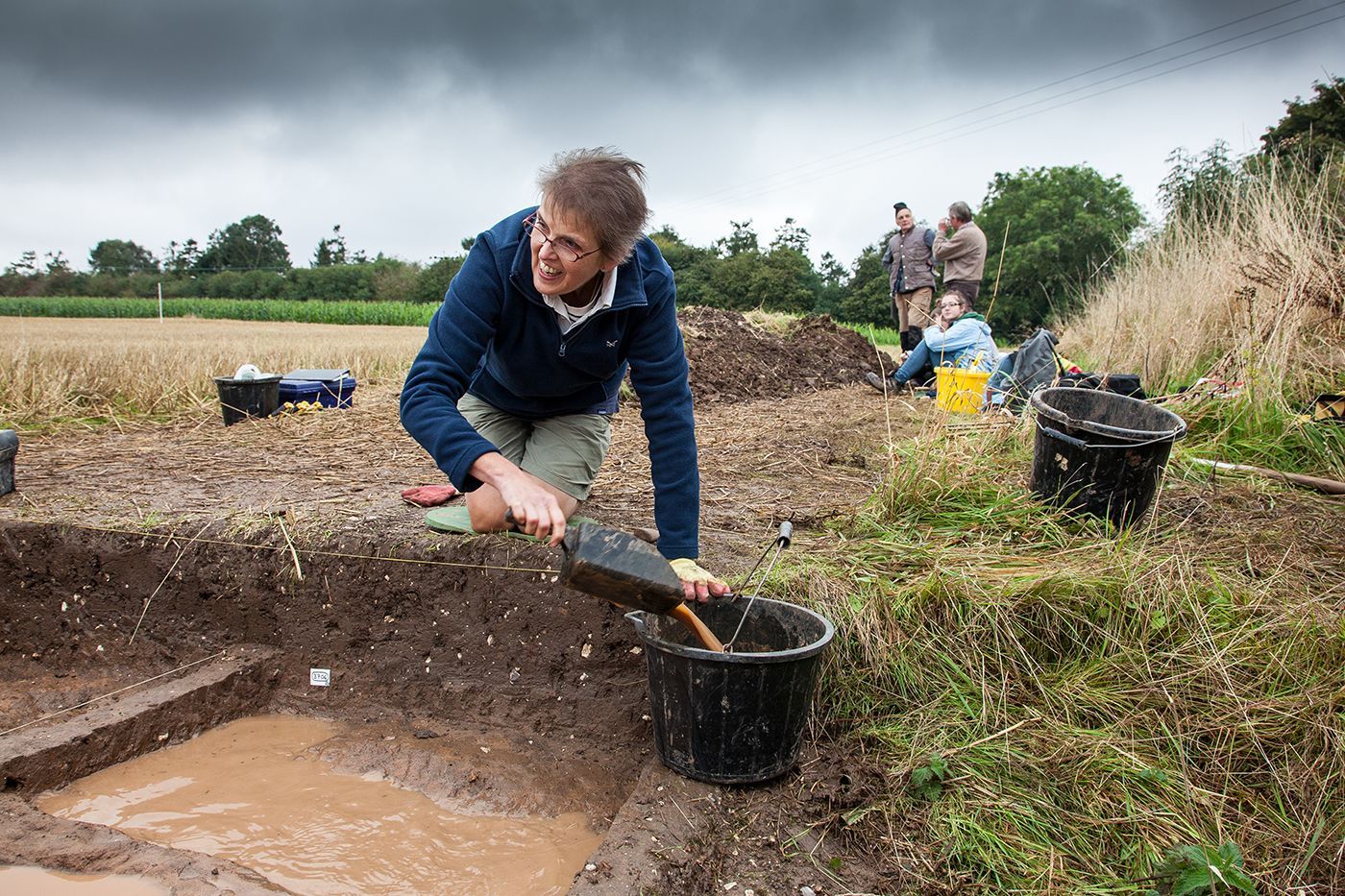 A woman is kneeling down in a field holding a shovel and pouring dirt into a bucket.