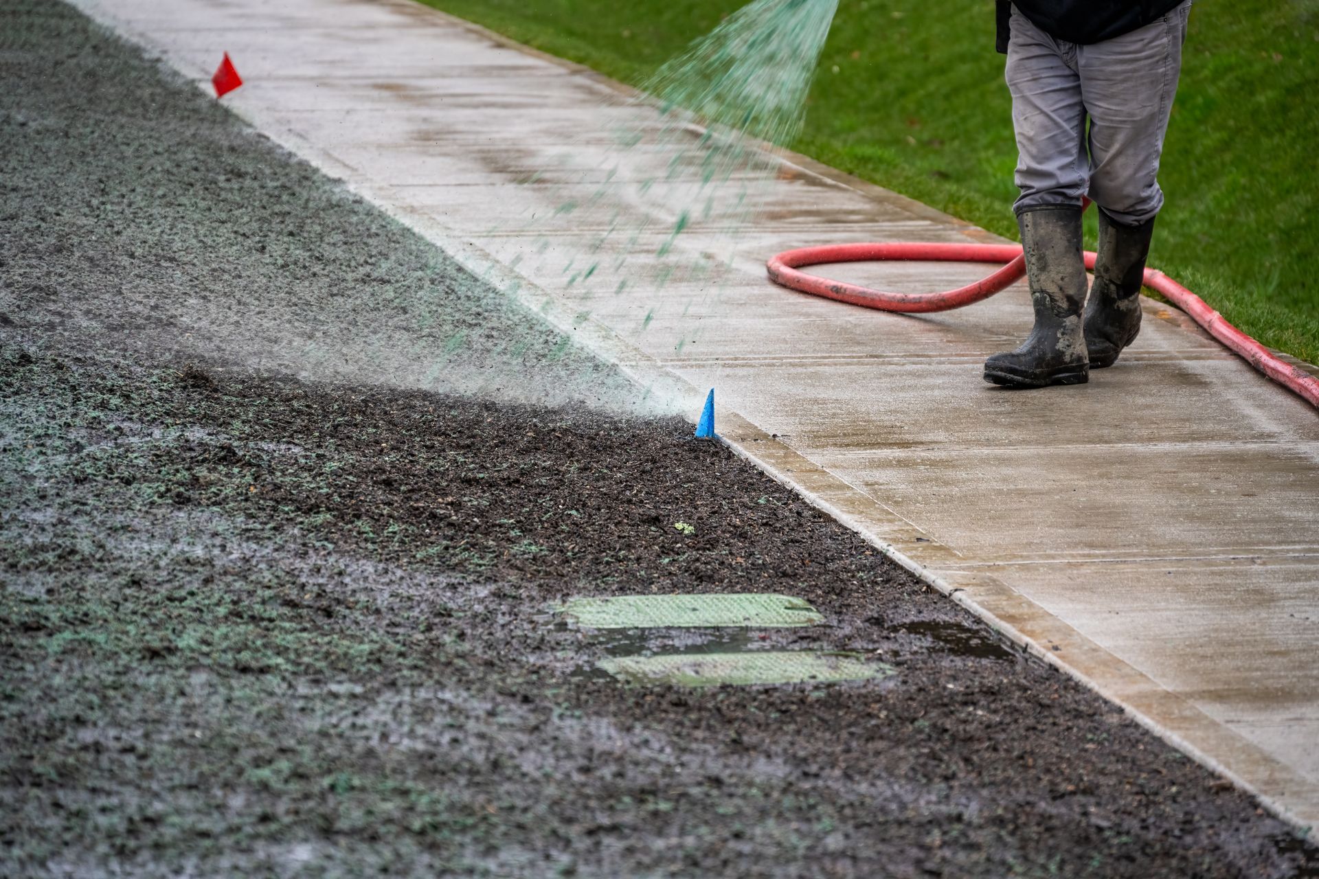 A person in boots uses a hose to spray water over a section of gravel next to a sidewalk and green grass.