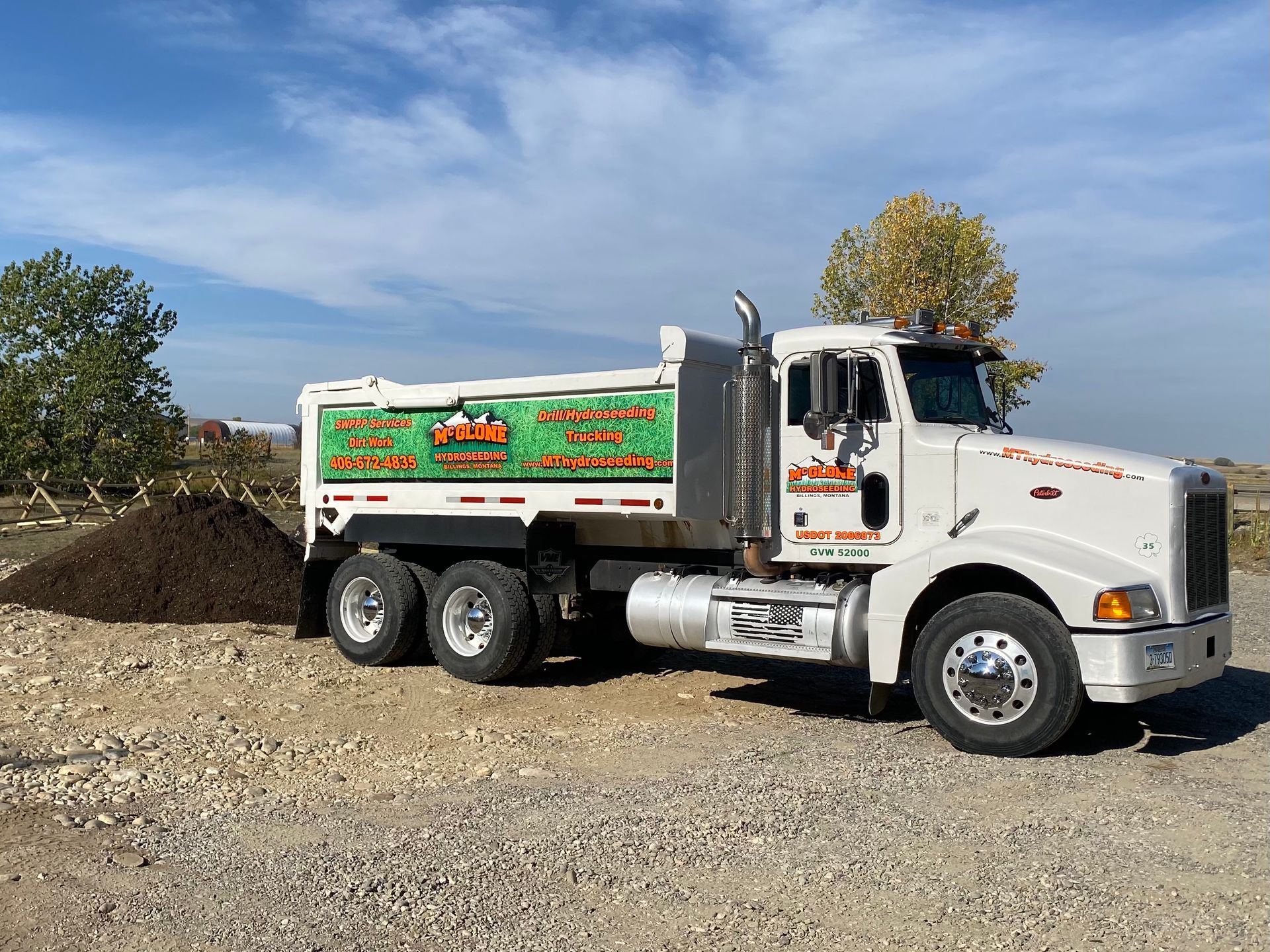 A white dump truck parked next to a pile of dark soil on a gravel lot under a clear blue sky.