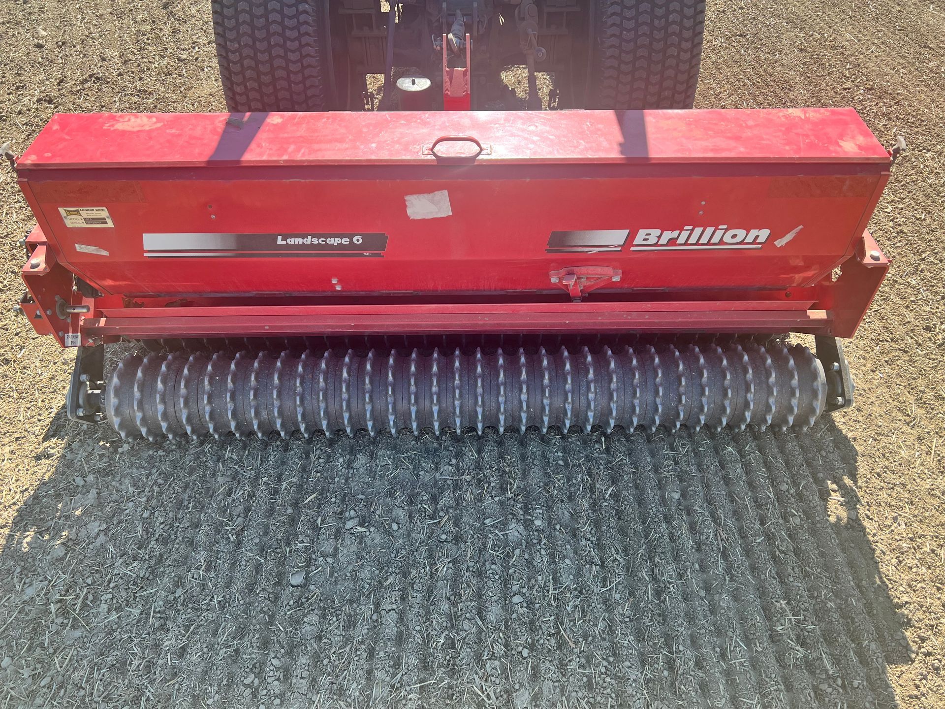 A red Brillion overseeder attached to a tractor, parked on a field of freshly prepared soil.