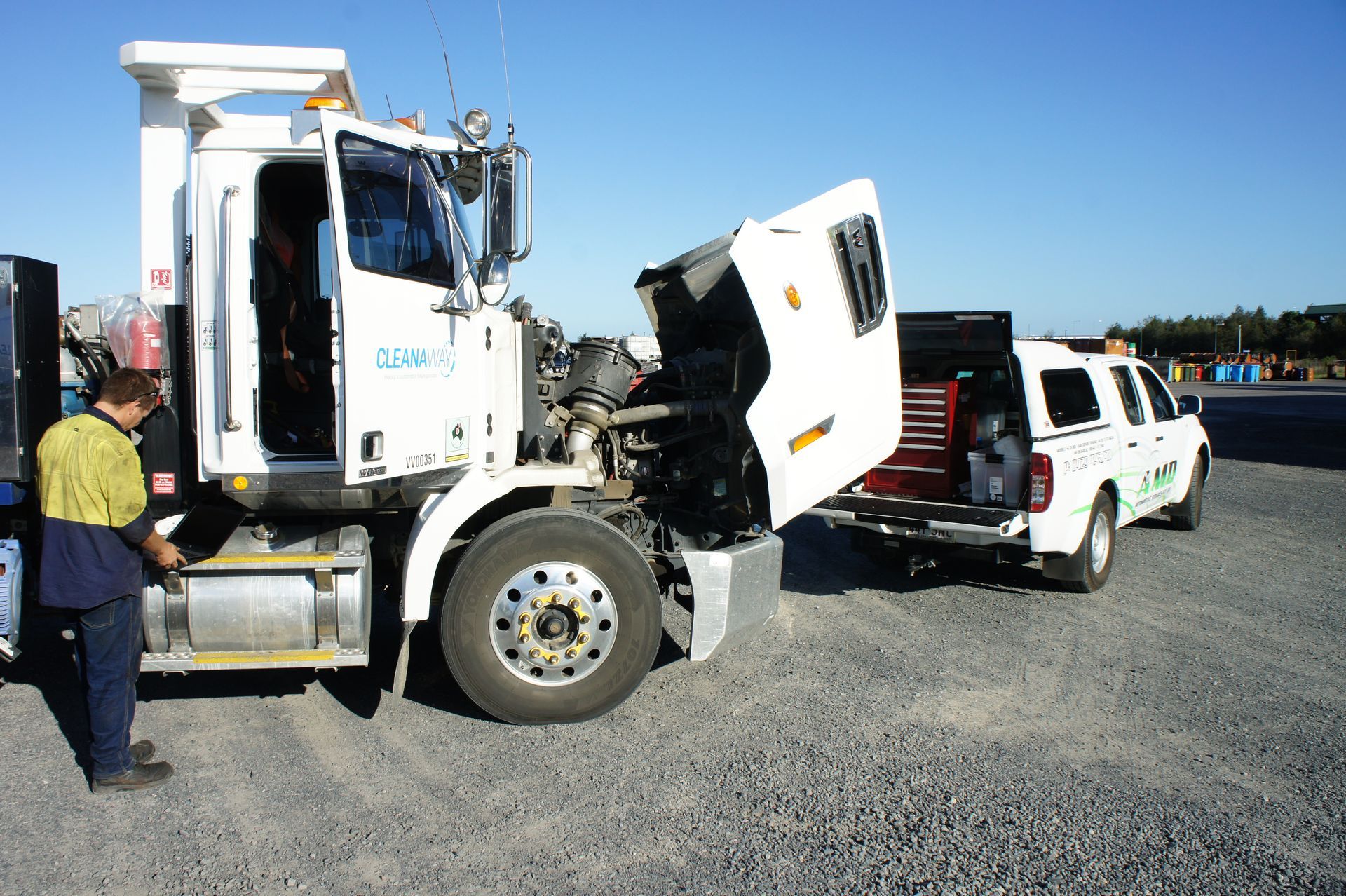 A mechanic standing by a heavy truck with its hood open for repairs, with a service vehicle parked behind it.