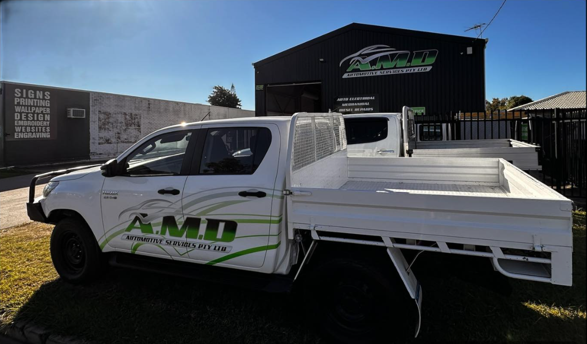 A white A.M.D. branded pickup truck parked in front of a matching company building on a sunny day.