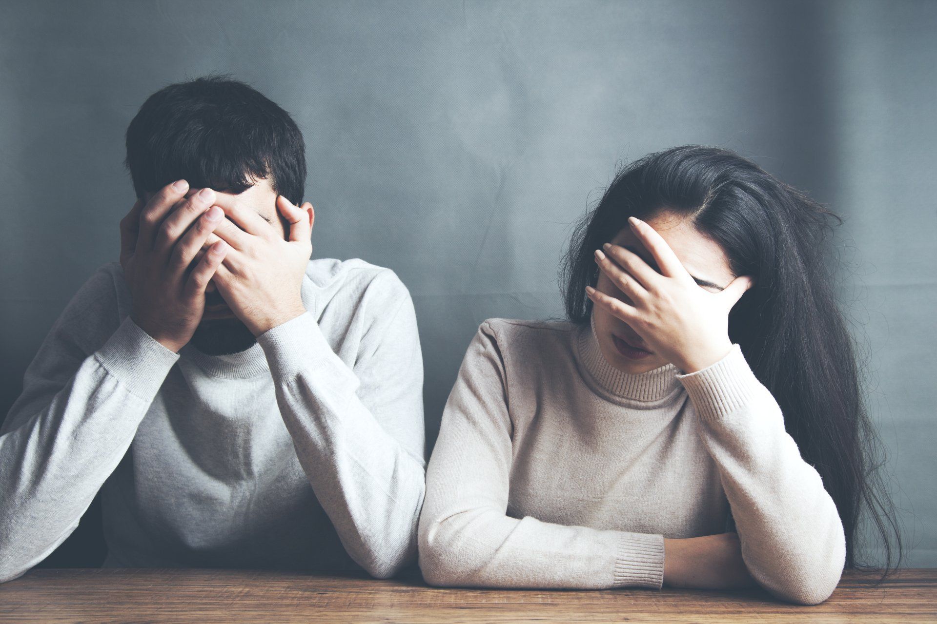 A man and a woman are sitting at a table covering their faces with their hands.