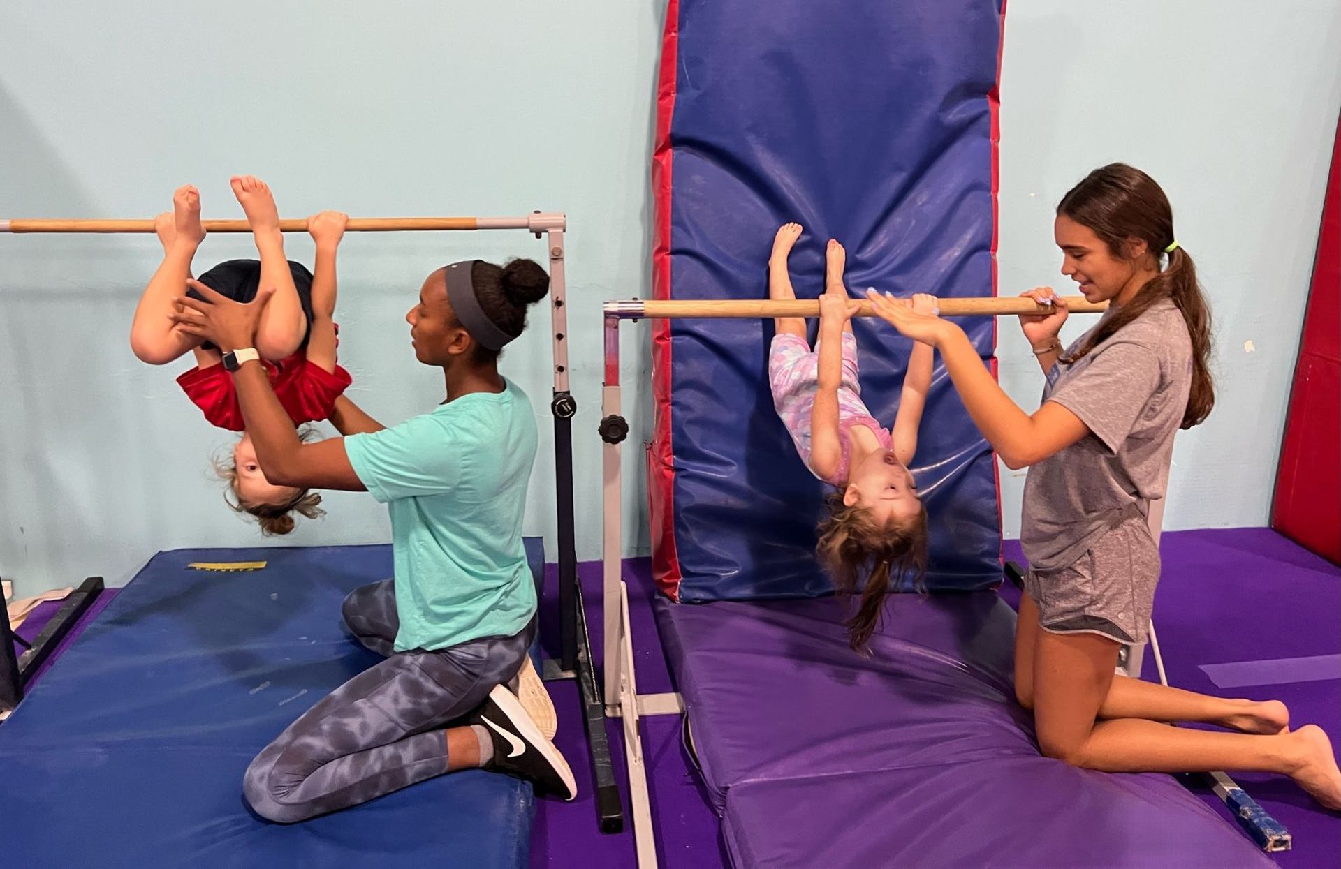 A woman is helping a child do a handstand on a bar in a gym.