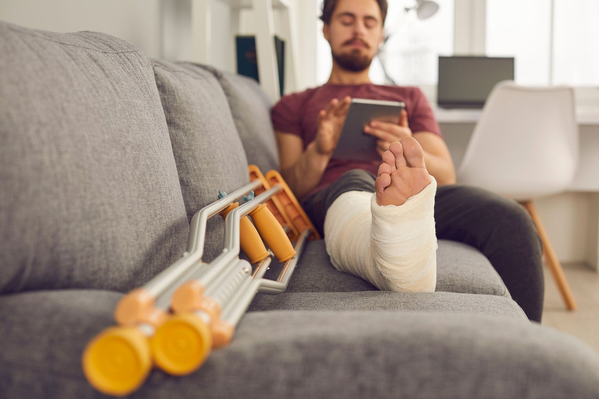 A person with a leg in a white cast sits on a grey sofa using a tablet, with crutches resting beside them.
