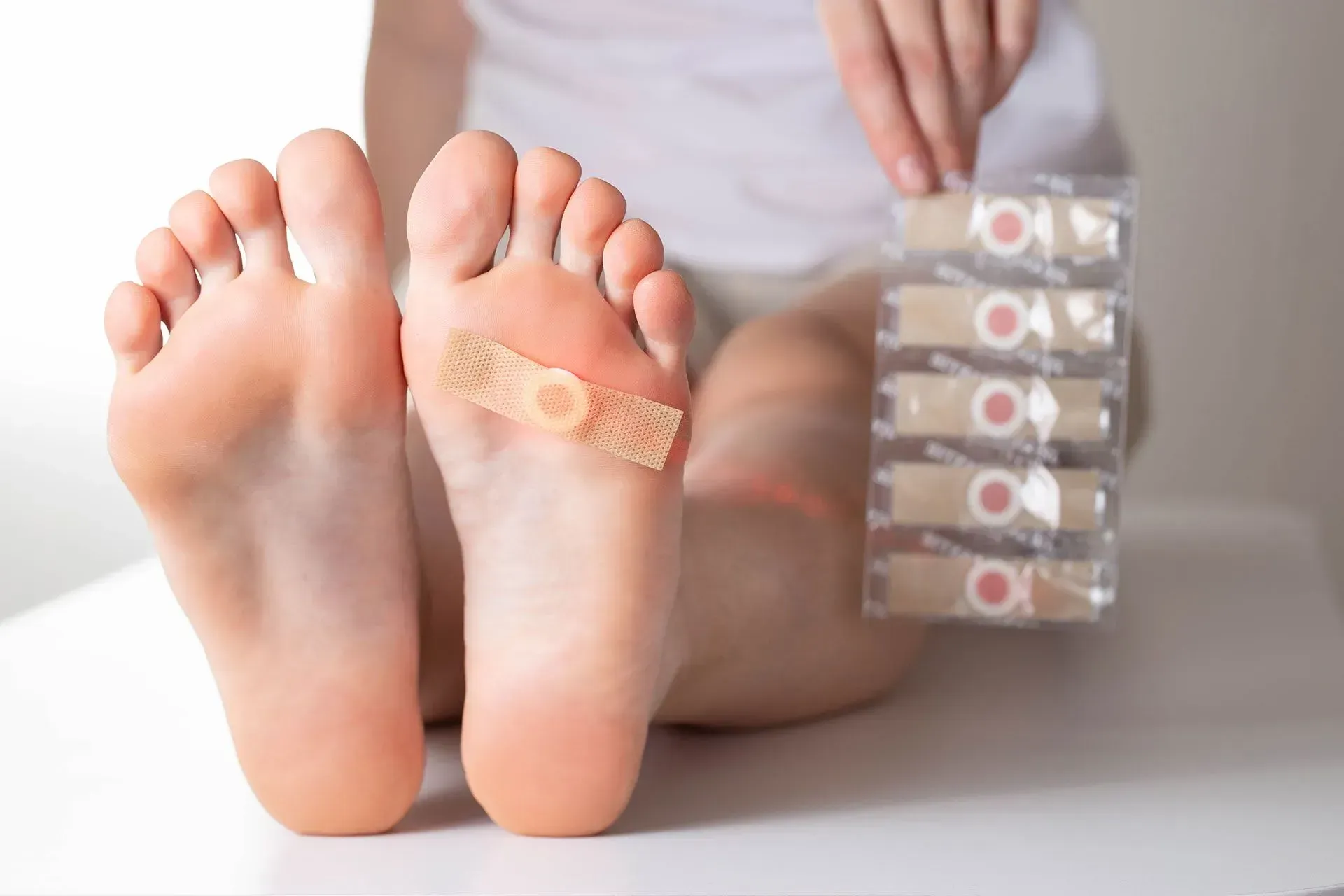 A person’s feet, one with a bandage on the sole, held near a pack of adhesive bandages.