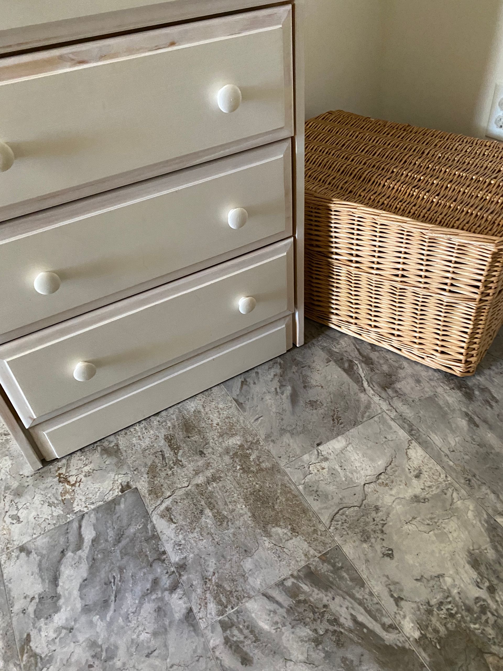 A wicker basket is sitting next to a dresser on a tiled floor.