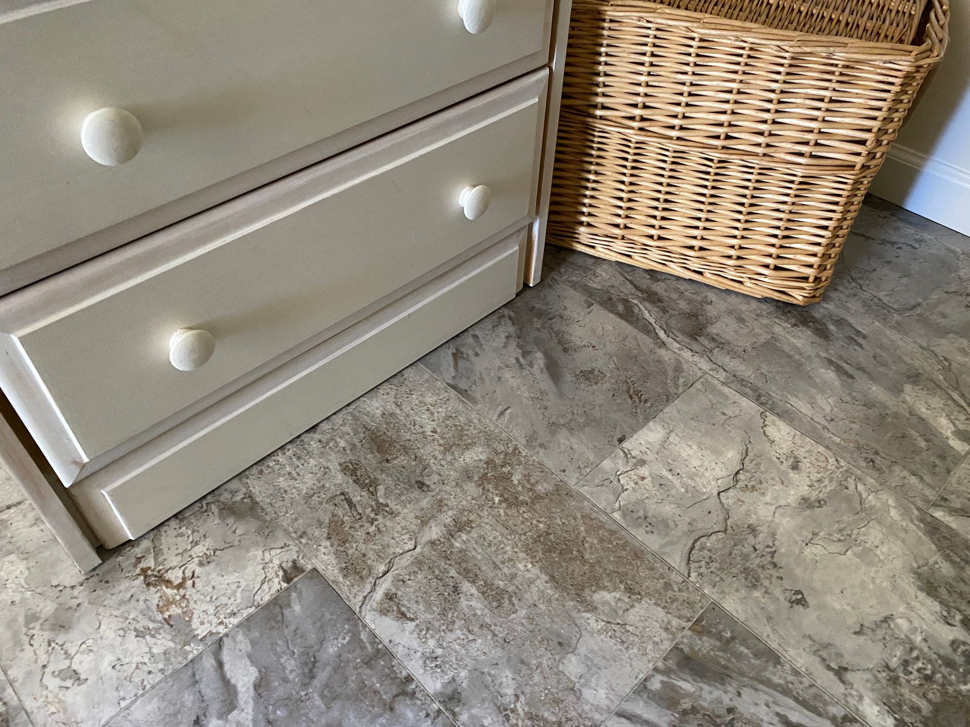 A wicker basket is sitting next to a dresser on a tiled floor.