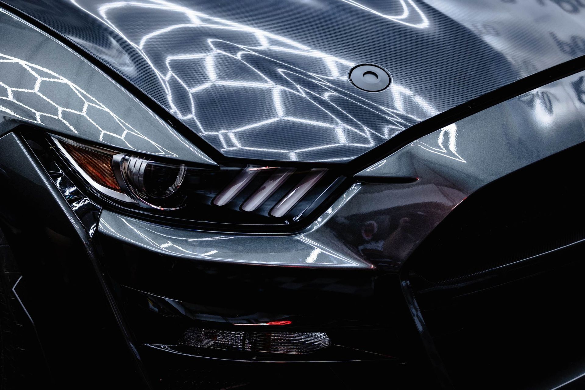 Close-up of a glossy, metallic dark grey Ford Mustang hood and headlight with a round hood pin in a well-lit workshop.