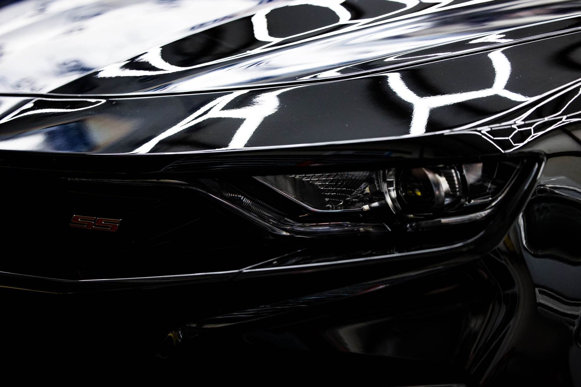 Close-up of a glossy black car's headlight and hood, reflecting bright workshop lights.