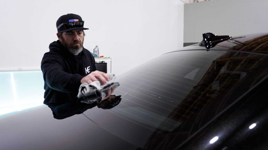 A person wearing a hat and hoodie wipes down the rear window of a black car in a white studio.
