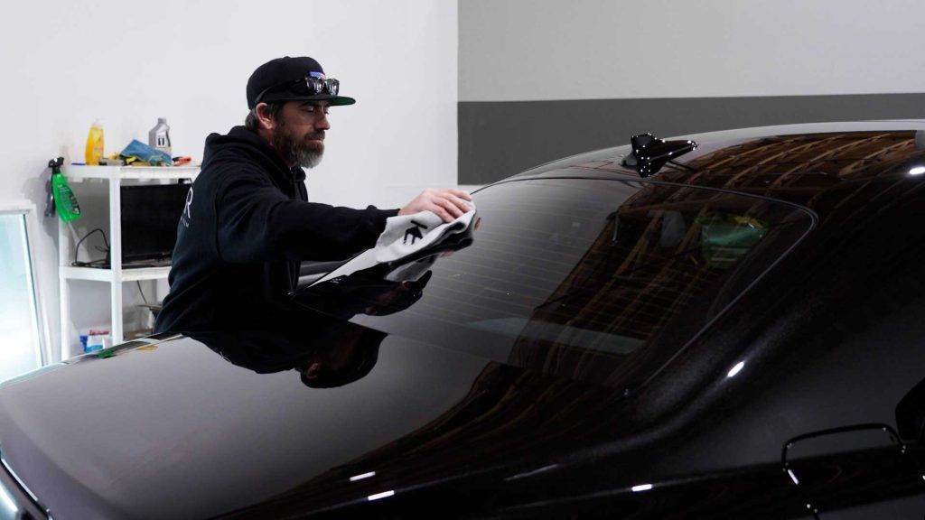 A person wearing a baseball cap and dark hoodie wipes the back window of a black car with a white cloth in a garage.