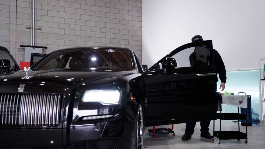 A person in dark clothing stands by the open door of a black Rolls-Royce inside a garage.
