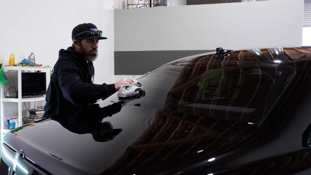 A person wearing a black cap and hoodie uses a polishing tool to buff the rear windshield of a black car in a garage.
