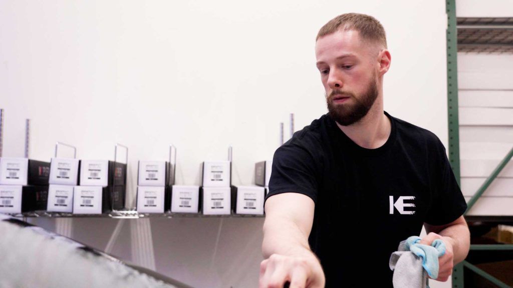 A person in a black t-shirt wipes a surface with a cloth in a room with shelves of labeled boxes in the background.