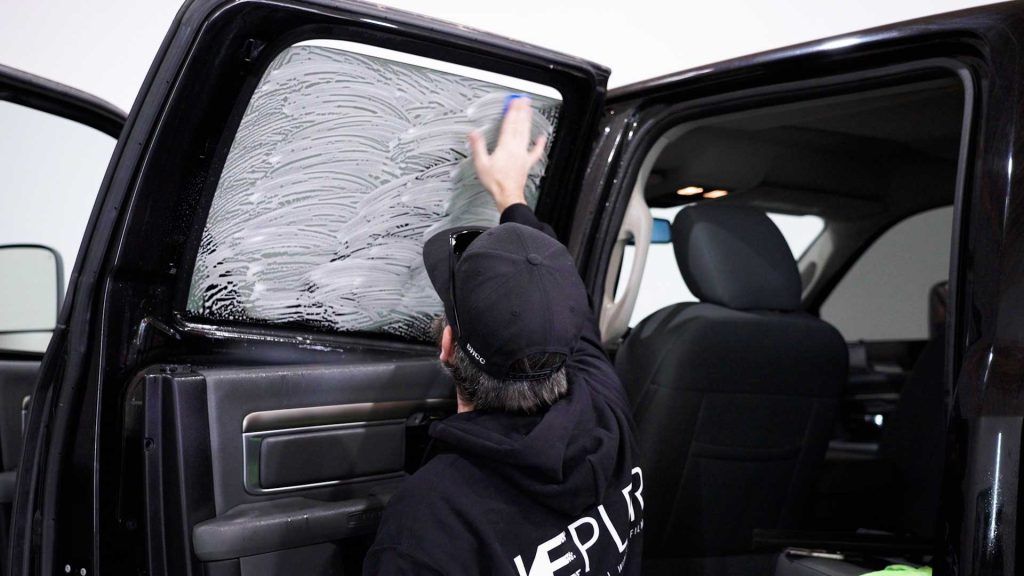 A person in a black hoodie applies a patterned, protective film to the interior side window of a black truck.