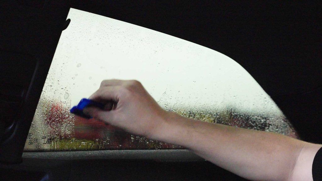 A hand uses a blue squeegee to clear water droplets from a vehicle's side window.