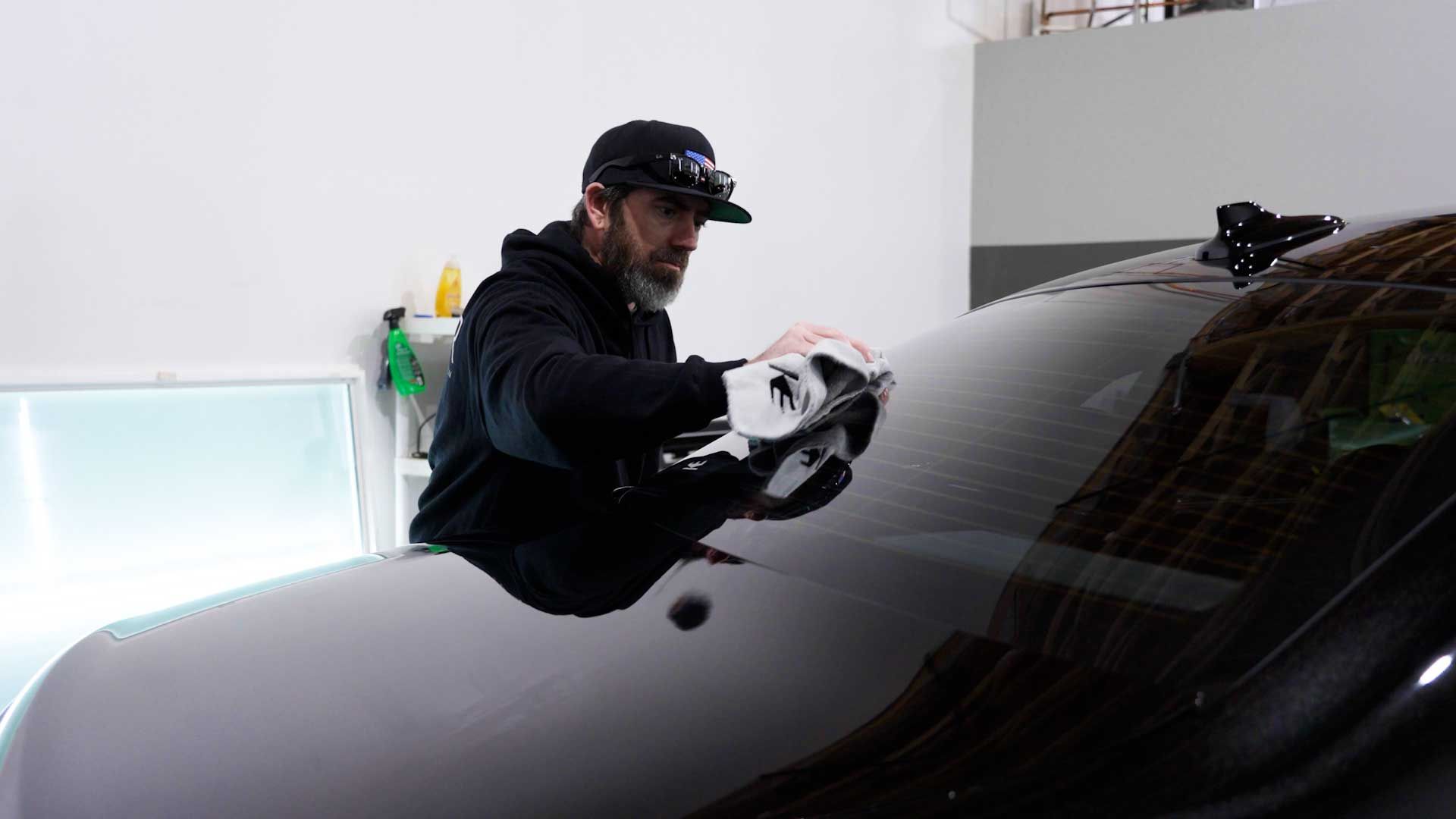 A person wearing a cap and hoodie wipes a dark car windshield with a white cloth inside a bright garage.