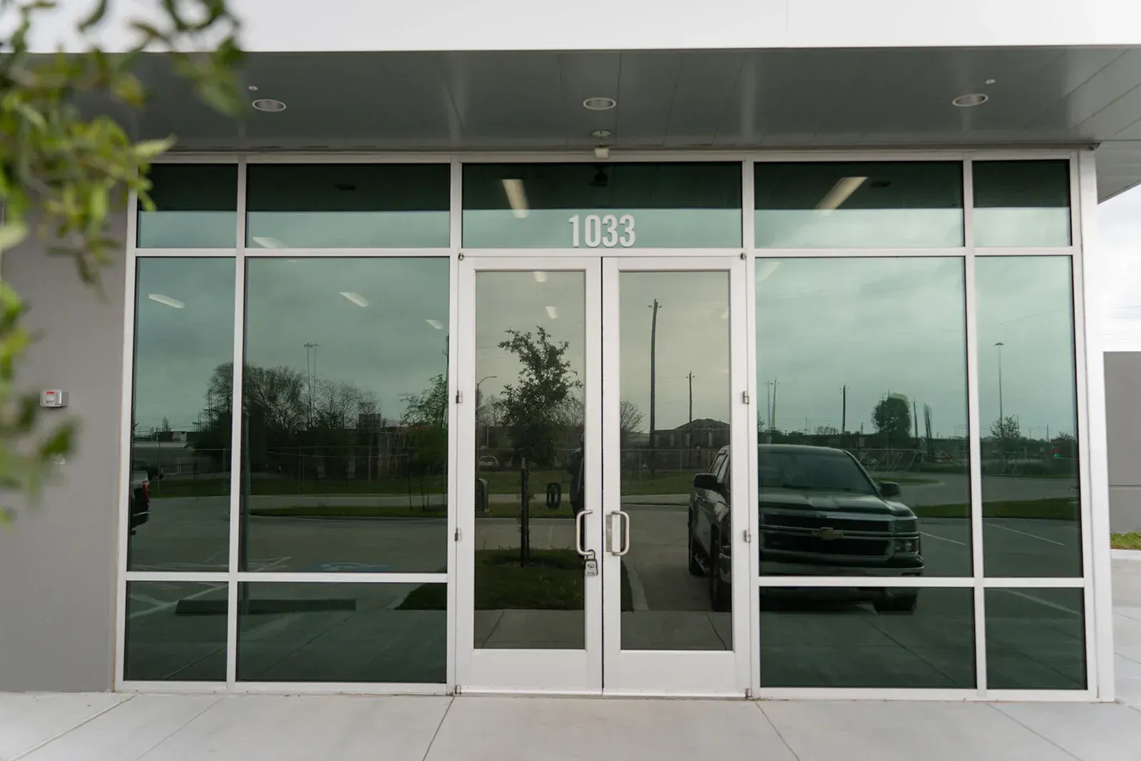 Glass-fronted commercial entrance with double doors, numbered 1033, reflecting a parking lot and vehicle.