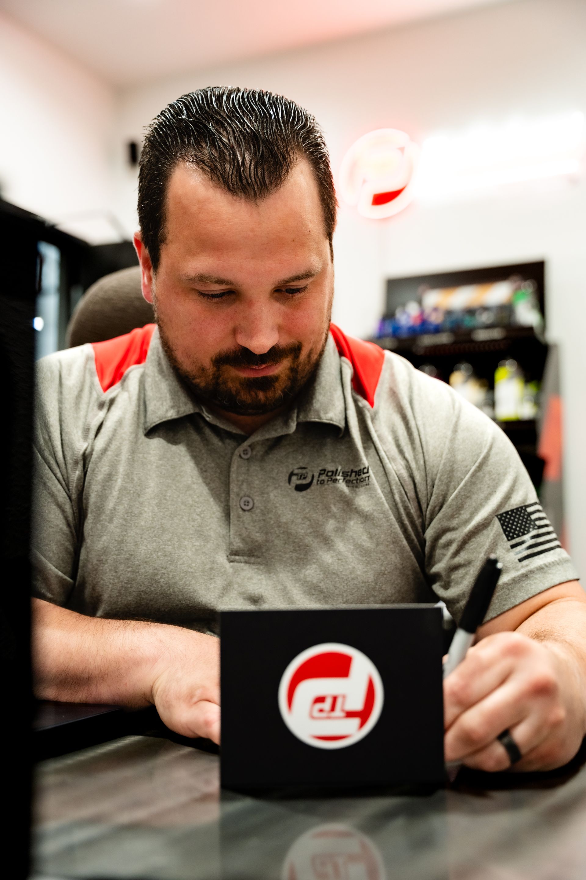 A person in a gray polo shirt signs an item with a red and white circular logo while seated at a desk.