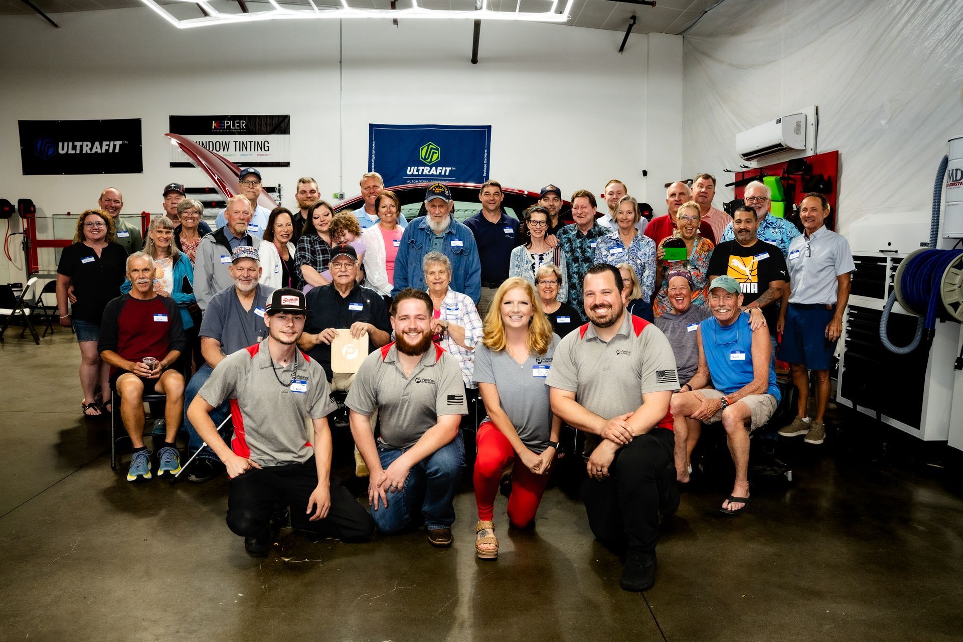 A large group of people smiling for a photo inside an automotive workshop with bright overhead lighting.
