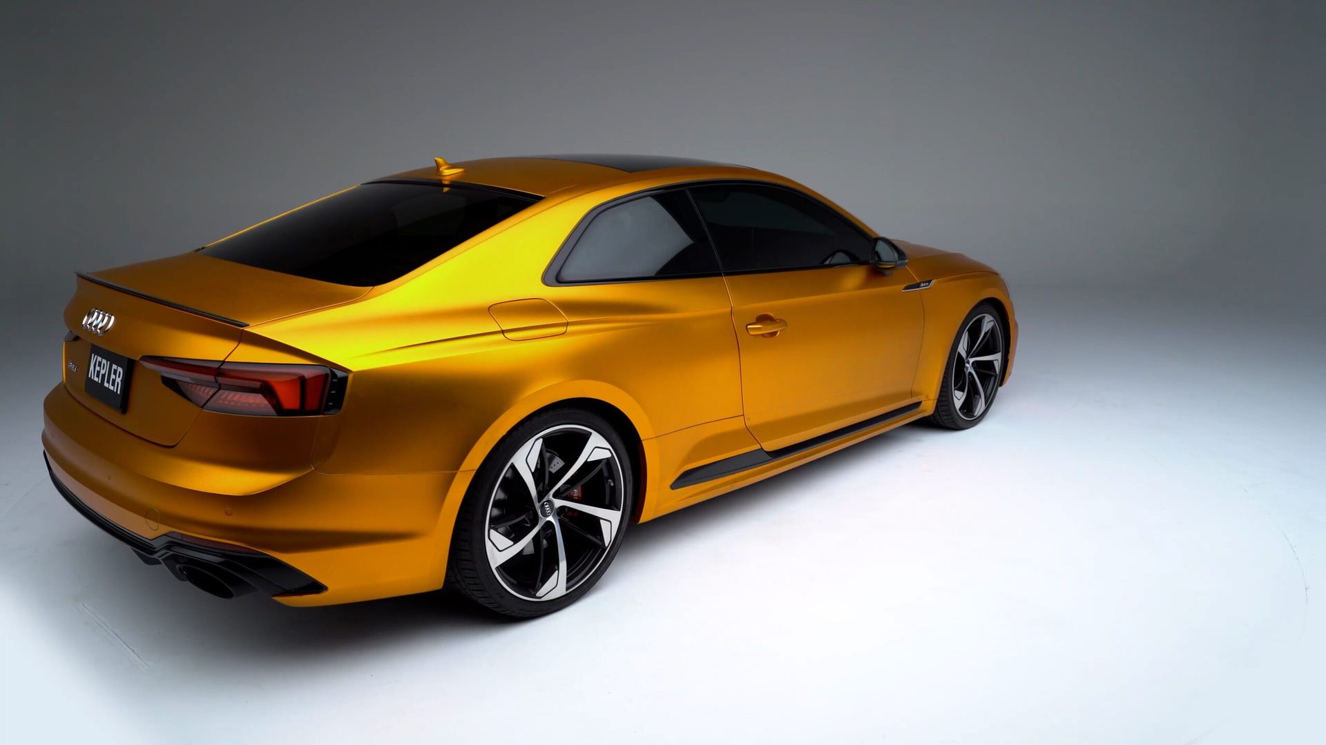 A metallic gold Audi S5 coupe viewed from the rear three-quarter angle against a neutral grey studio background.