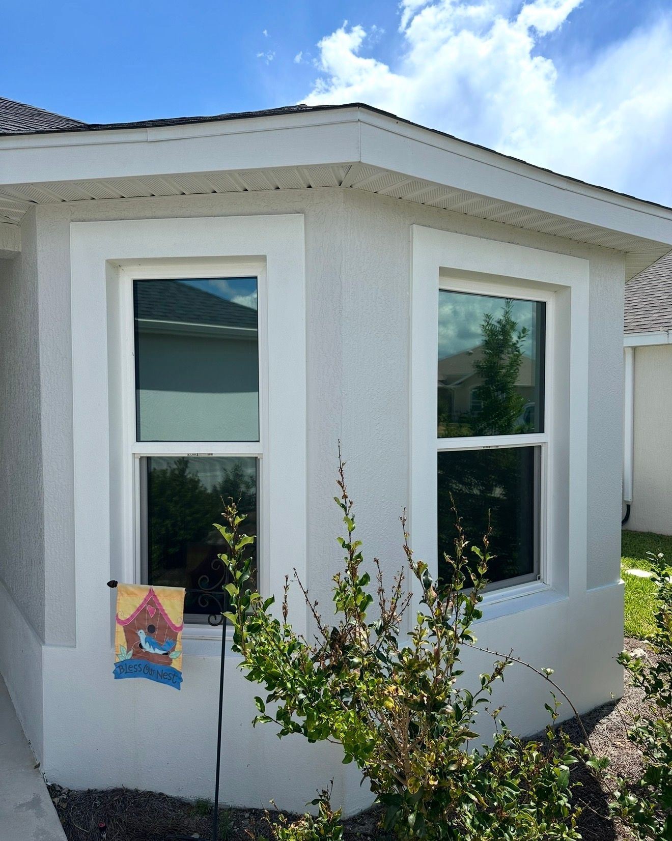 Two windows with white trim on a light-colored stucco house exterior, with a small decorative flag hanging on the left.