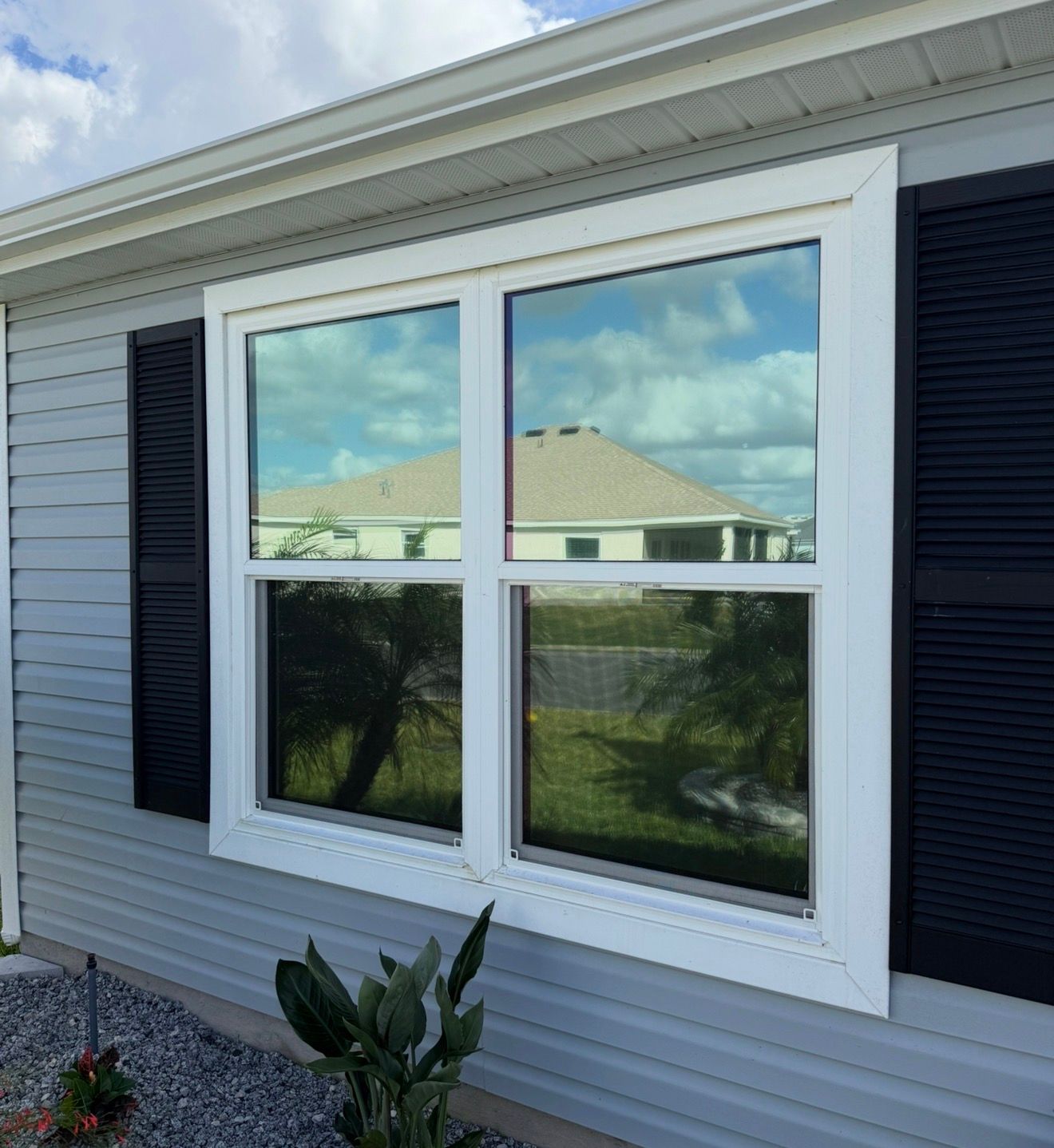 A white-framed double-hung window on a light gray vinyl-sided house with black shutters reflecting a neighboring house.