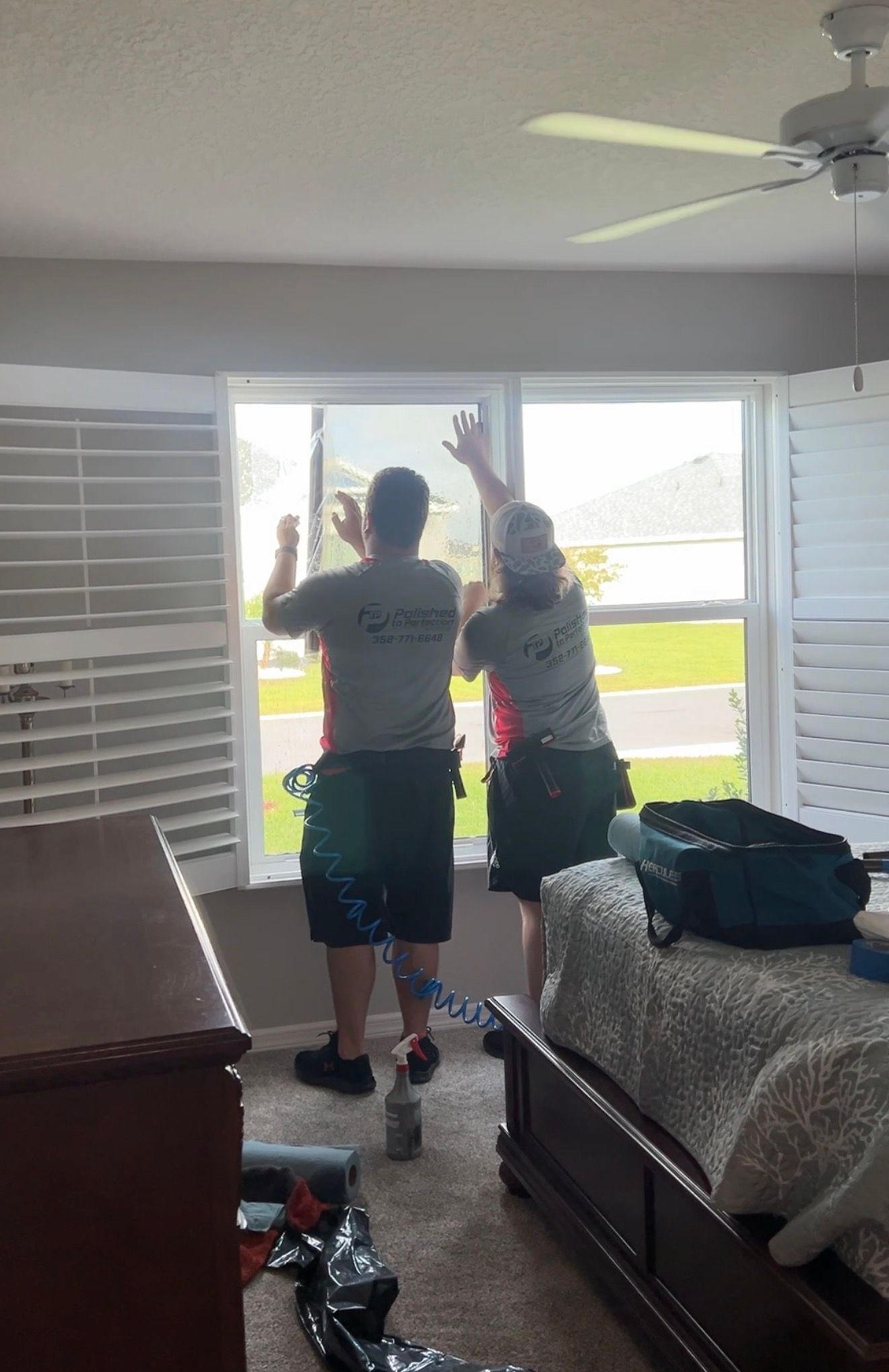 Two people in matching gray shirts and dark shorts apply window tint film to a home window in a bedroom.