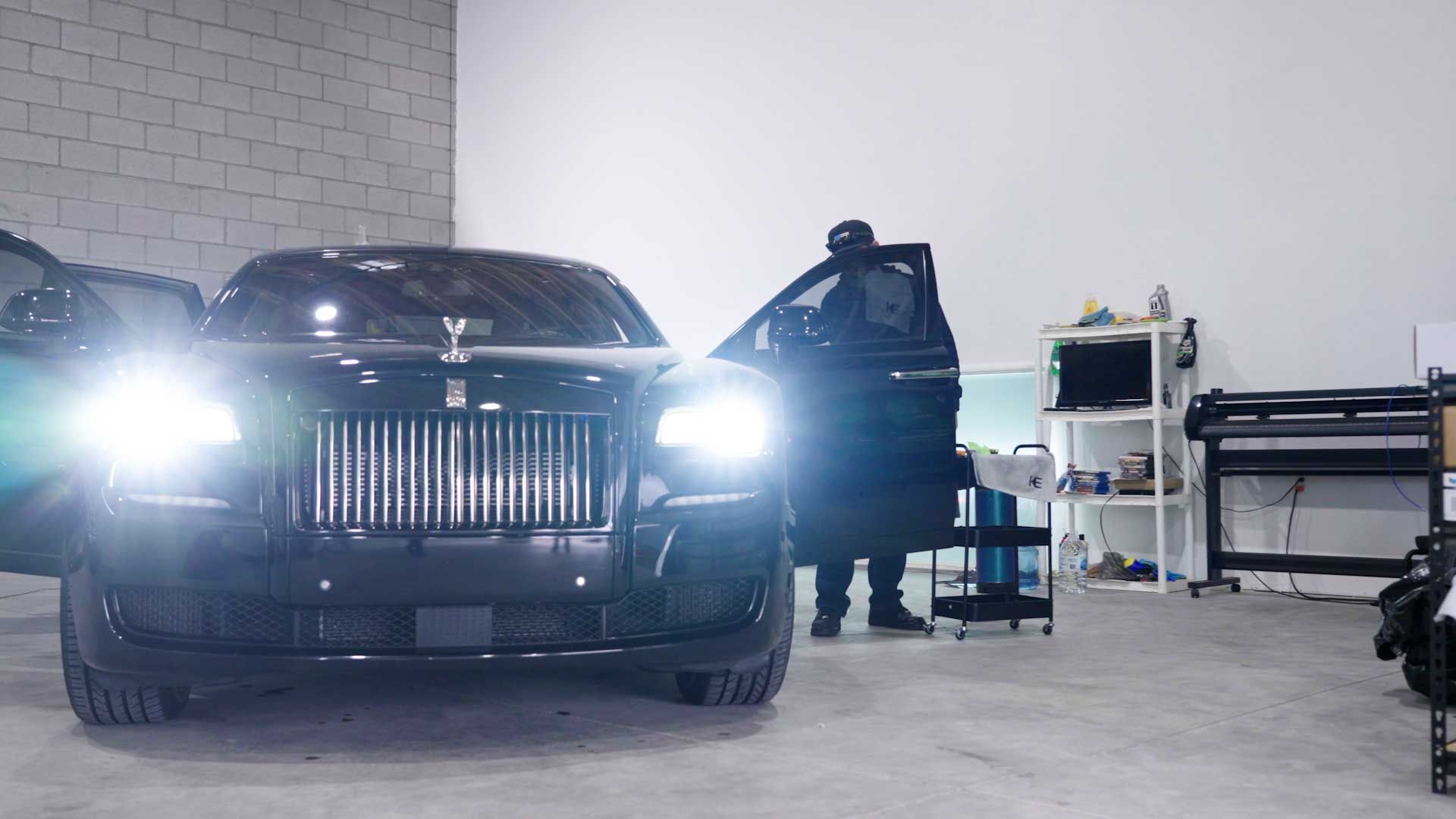A black Rolls-Royce with its headlights on is parked inside a garage, with a person standing by the open driver-side door.