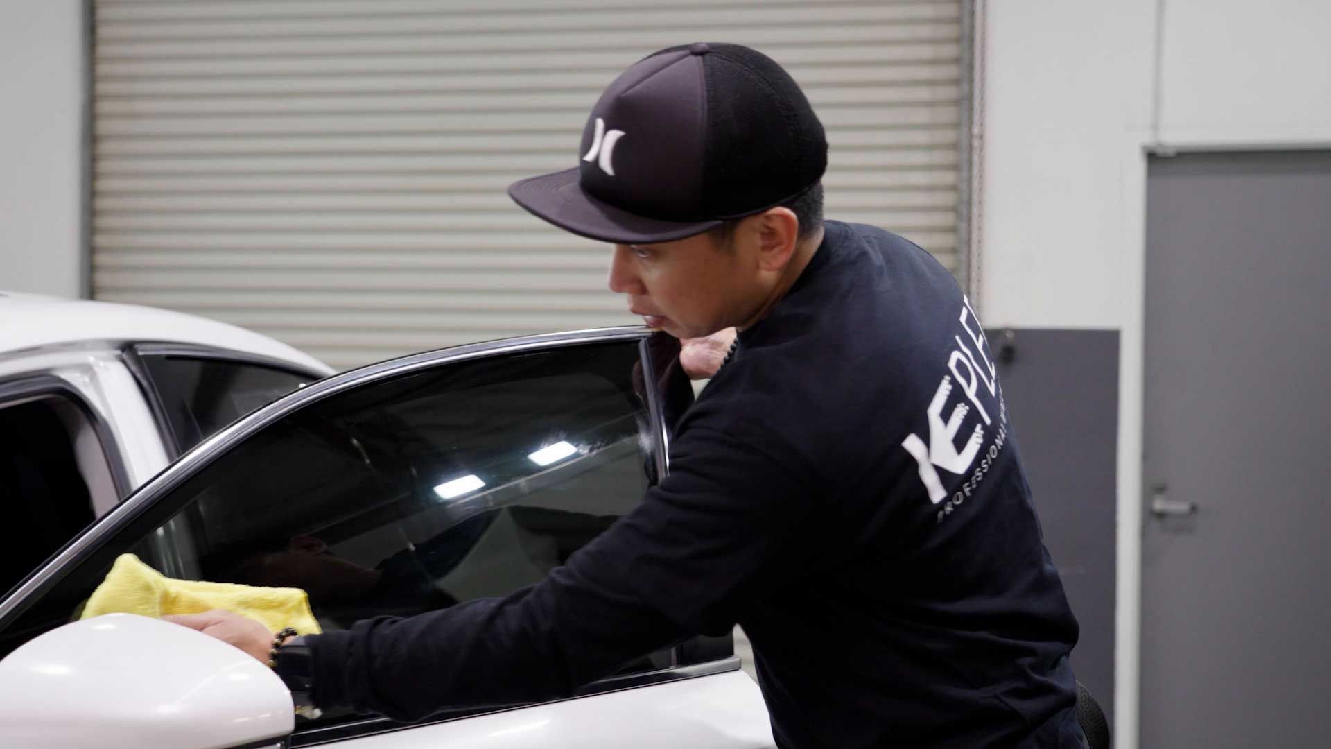 A person wearing a dark shirt and hat uses a yellow cloth to clean the side window of a white car in a garage.