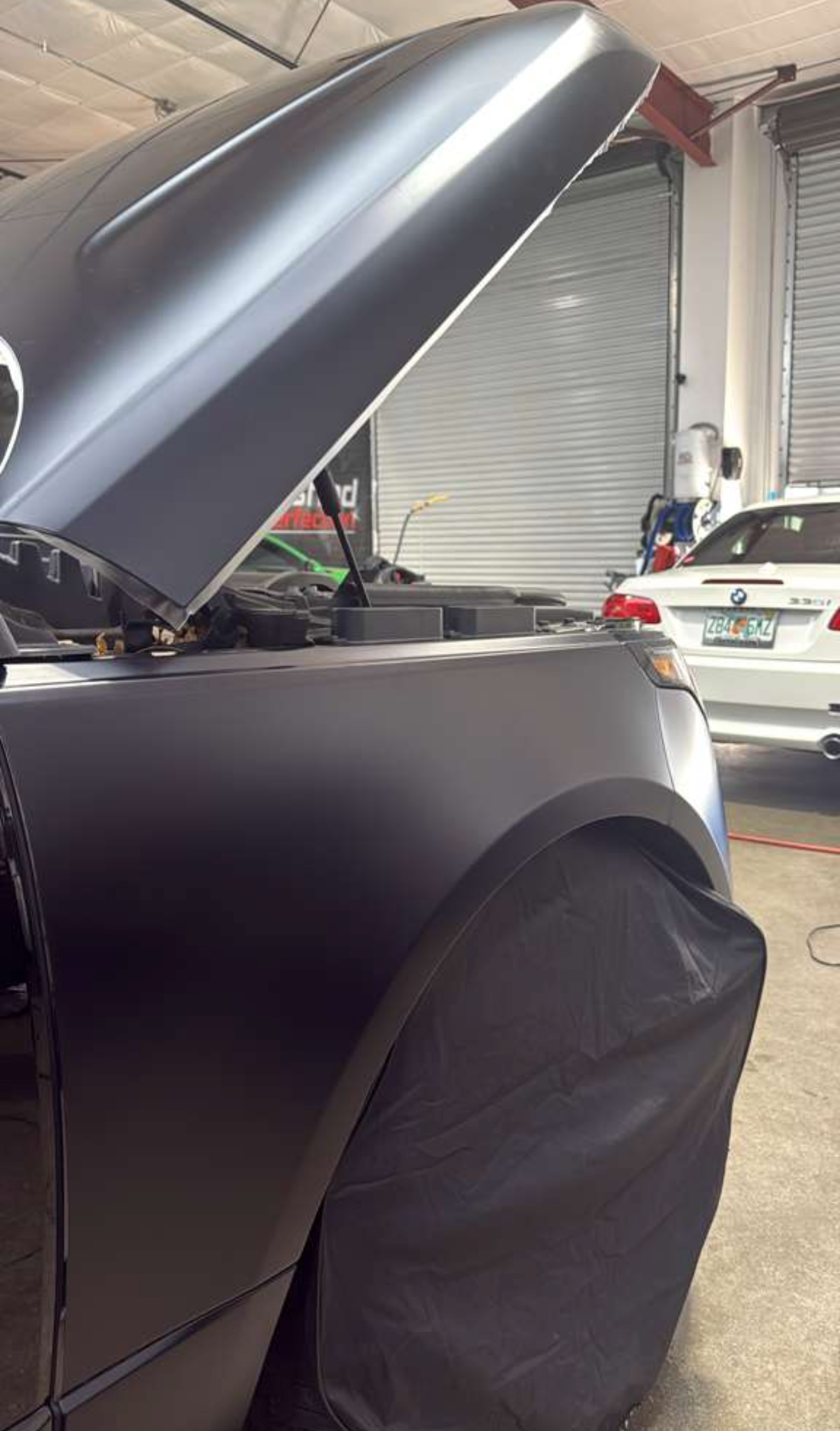 A matte black car with its hood open in a workshop, with a protective cover over the front wheel.