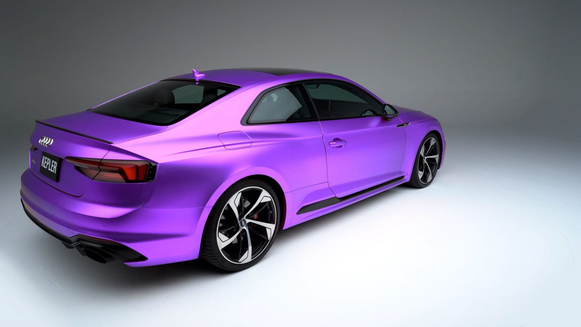 A sleek, matte purple sports coupe shown from the rear three-quarter angle in a minimalist studio setting.