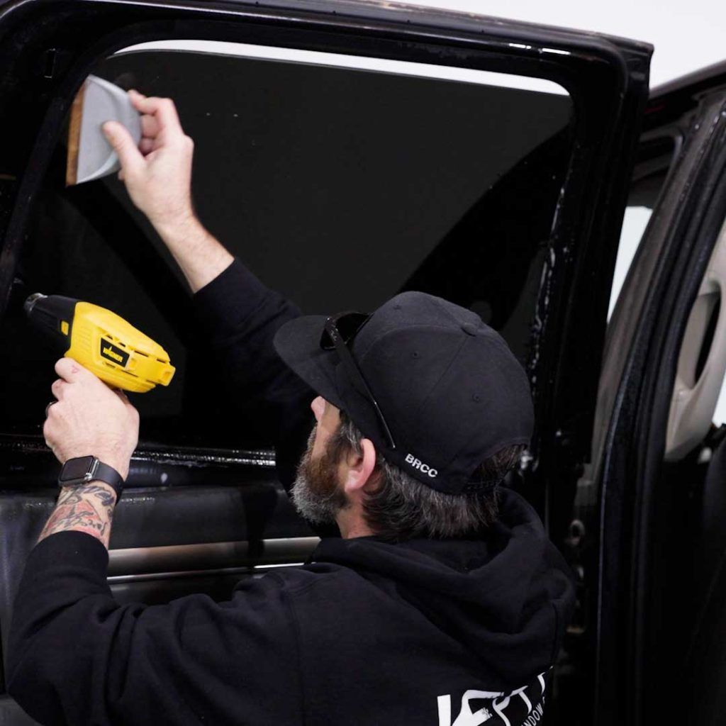 A person wearing a black cap and hoodie uses a heat gun and a squeegee to apply dark window tint to a car door window.
