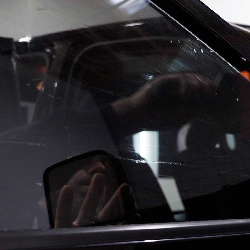 A hand applying a clear film to a car window, with the reflection of the person visible in the side-view mirror.