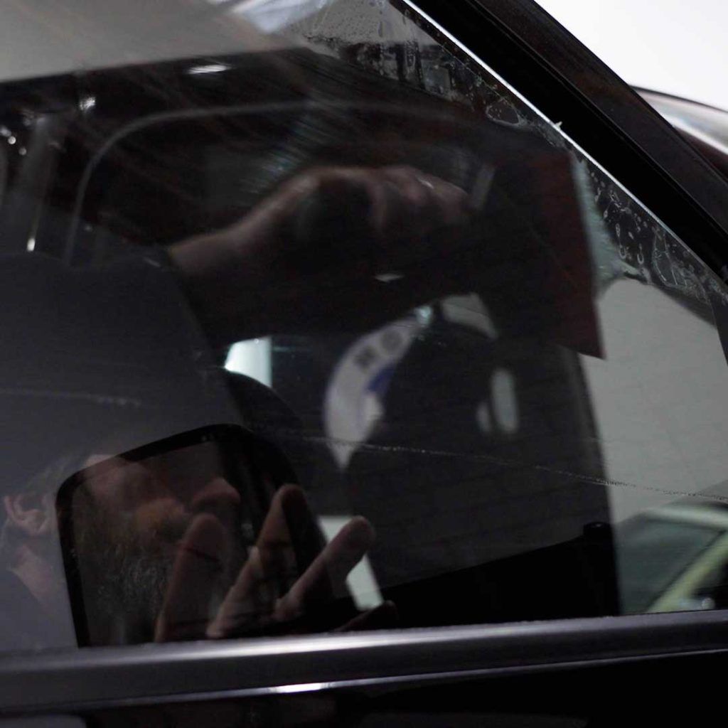 A person uses a squeegee to smooth window tint film onto a car window.