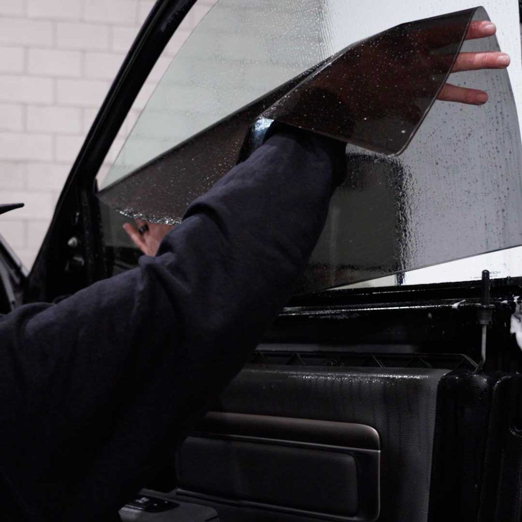 A person’s hand applies a tinted film to a wet car window.