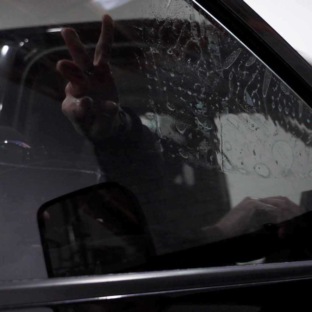 A person seen through a car window makes a peace sign with their hand against the glass, which is covered in water droplets.