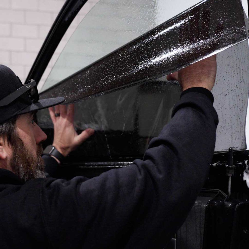 A person in a black cap and hoodie applies a layer of wet window tint film to a car door window.