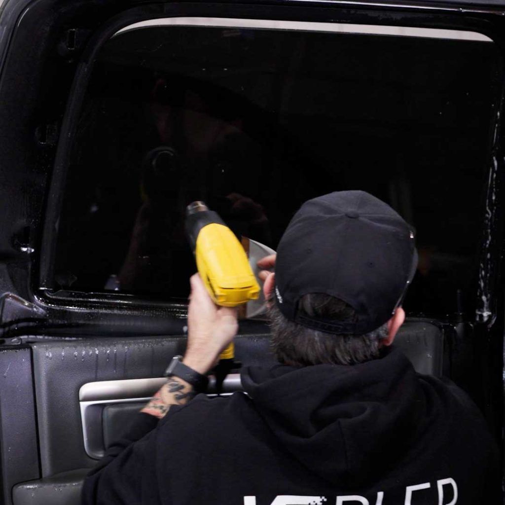 A person in a black hoodie and cap uses a yellow heat gun to apply window tint to a car door window.